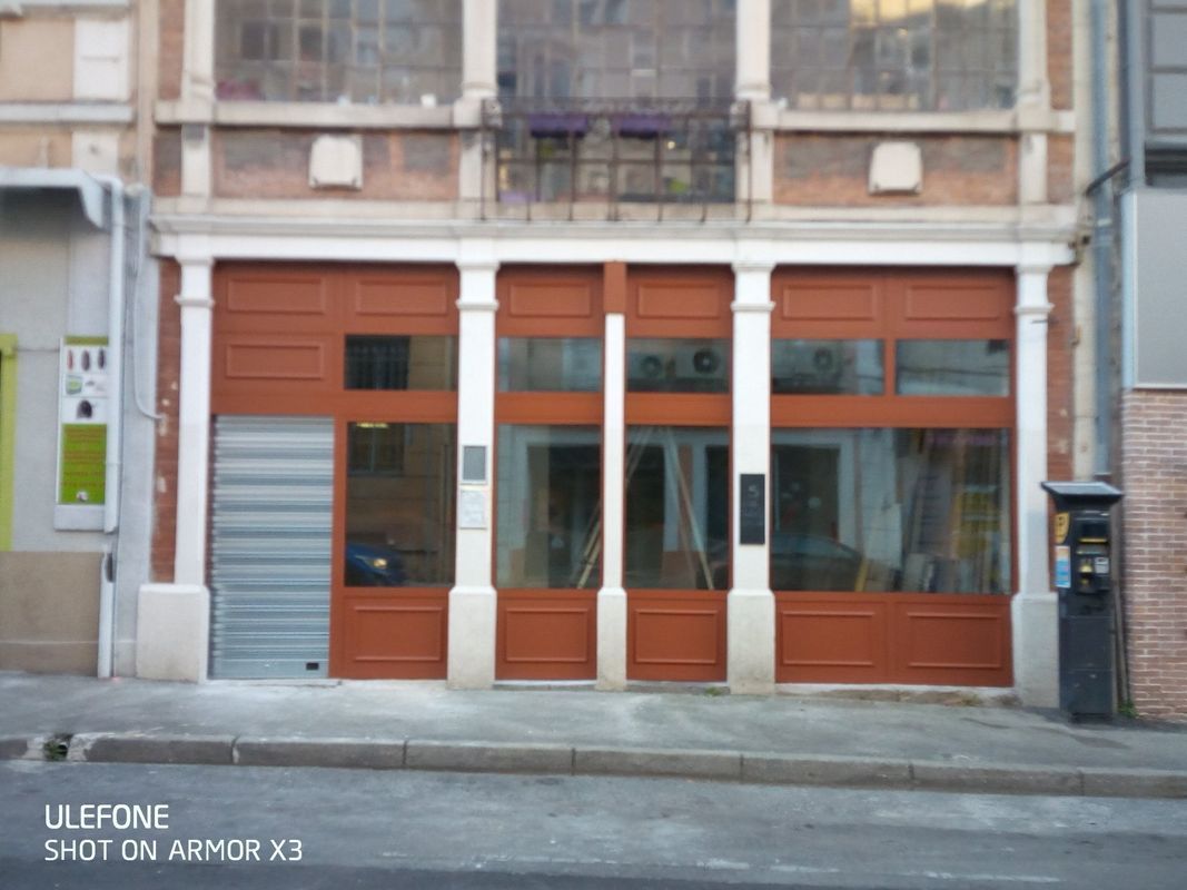 Une façade de magasin avec un encadrement en bois brun rougeâtre et de grandes fenêtres, présentant un volet roulant de sécurité métallique fermé sur la gauche.