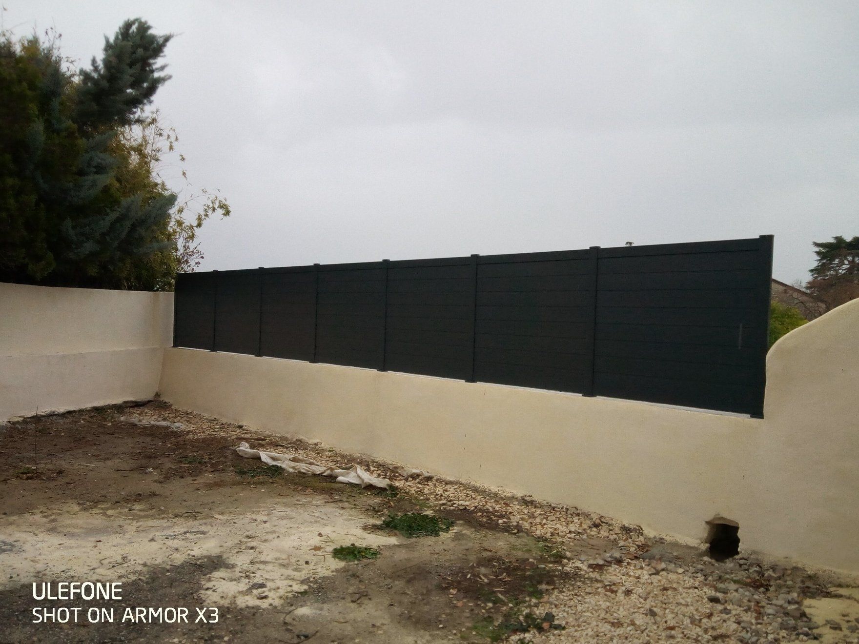 Des panneaux d'intimité noirs sont fixés au sommet d'un muret en maçonnerie beige, dans un espace extérieur au sol en terre battue.