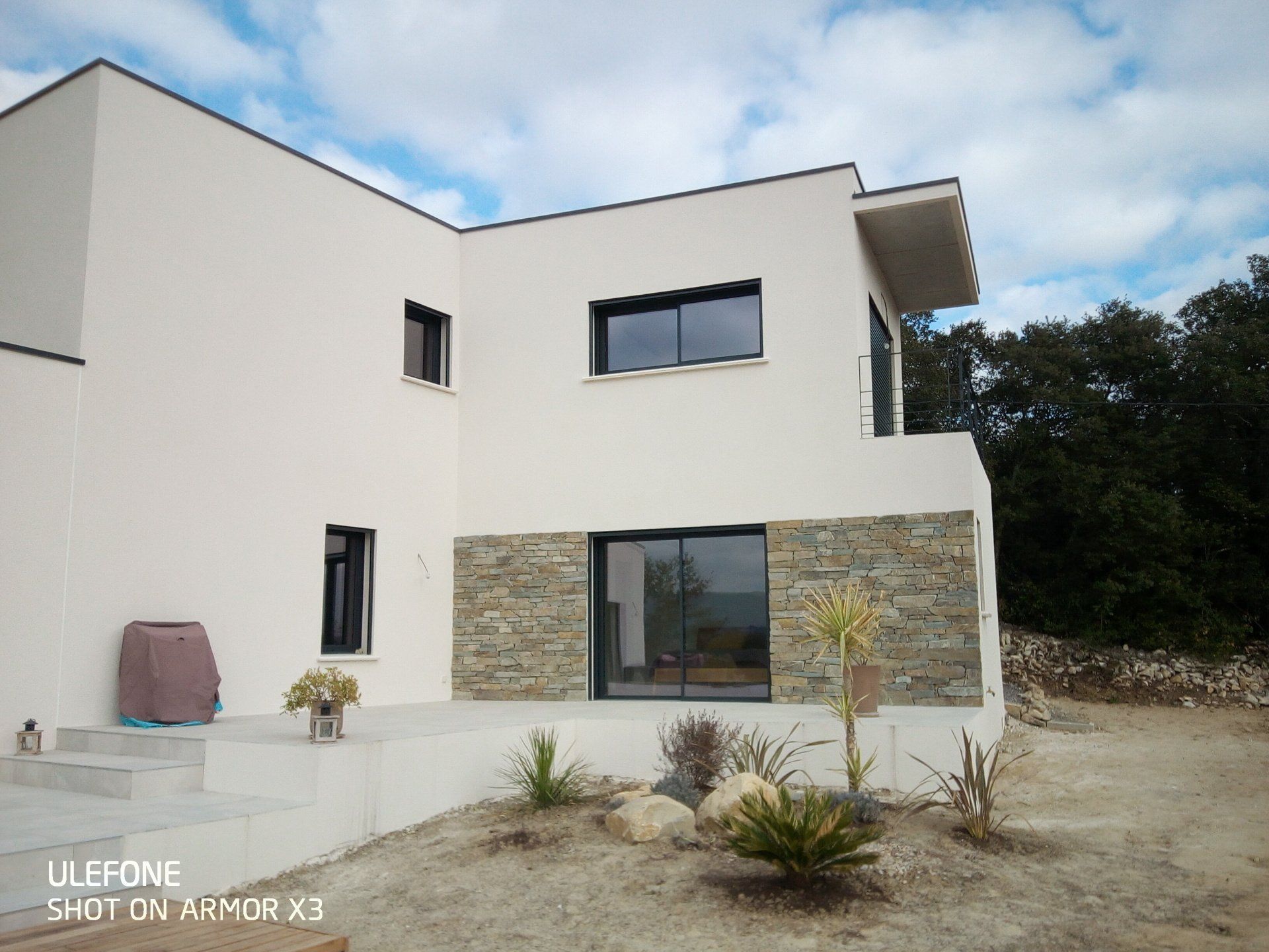 Une maison moderne de deux étages, aux tons clairs, avec un soubassement en pierre, de grandes fenêtres et une terrasse pavée.
