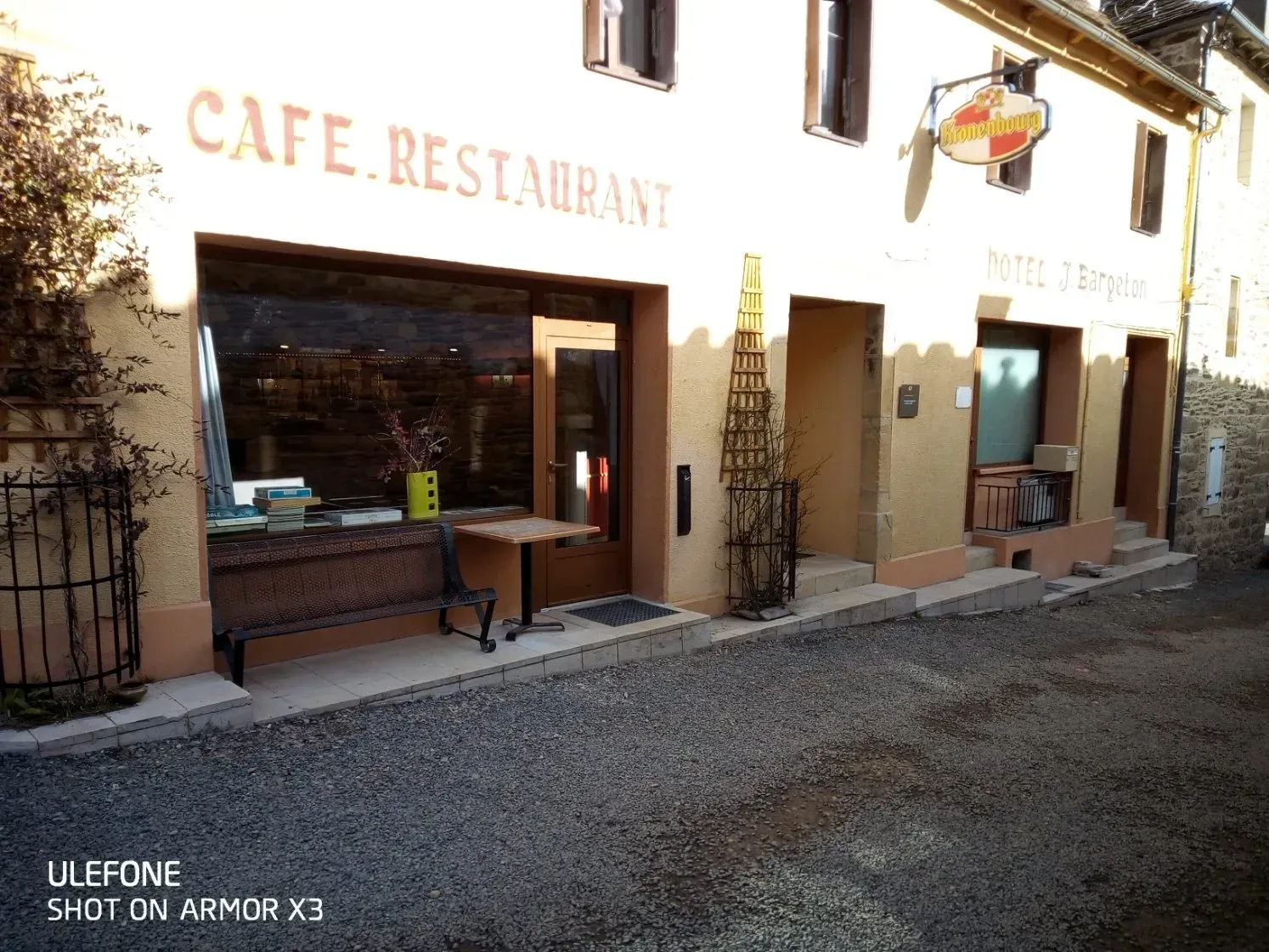 Un bâtiment de deux étages, de couleur beige, abritant un café-restaurant, avec un premier plan recouvert de gravier, un banc et une petite table devant l'entrée.