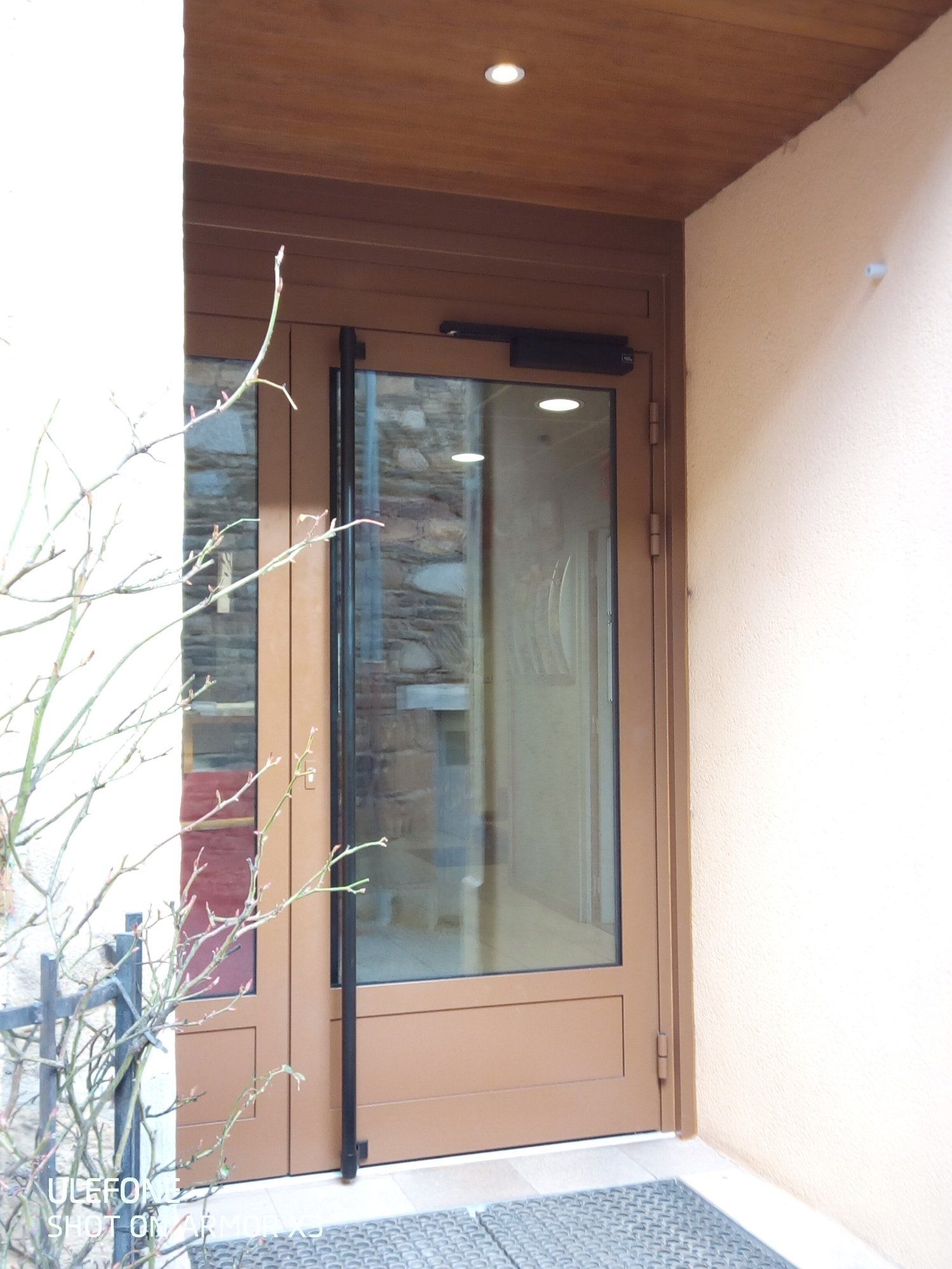 Une porte marron avec une grande poignée verticale noire et une fenêtre en verre, située dans une entrée en retrait d'un bâtiment.