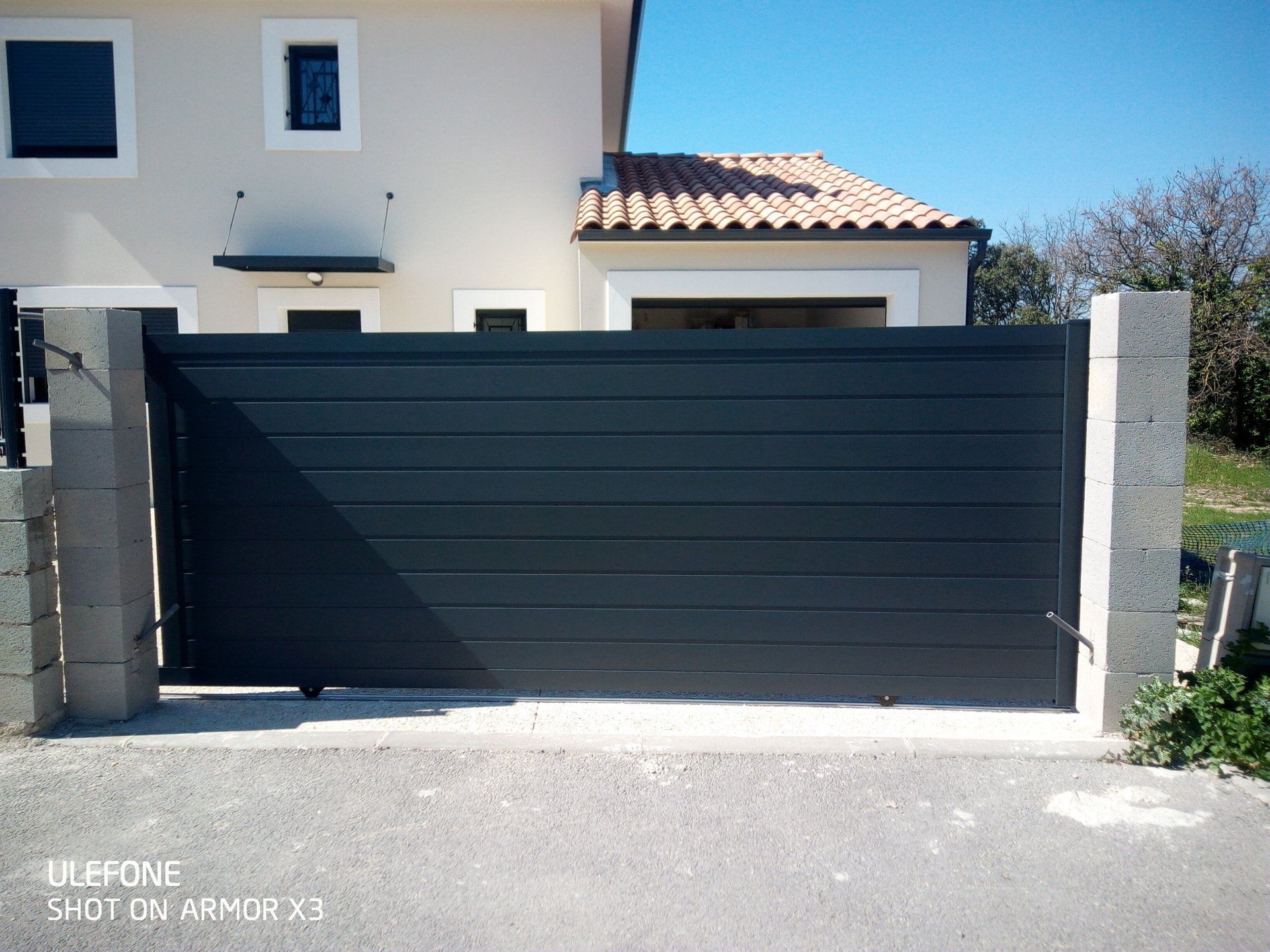 Un portail coulissant à lattes horizontales gris foncé, installé entre deux piliers de pierre devant une maison moderne de couleur claire.