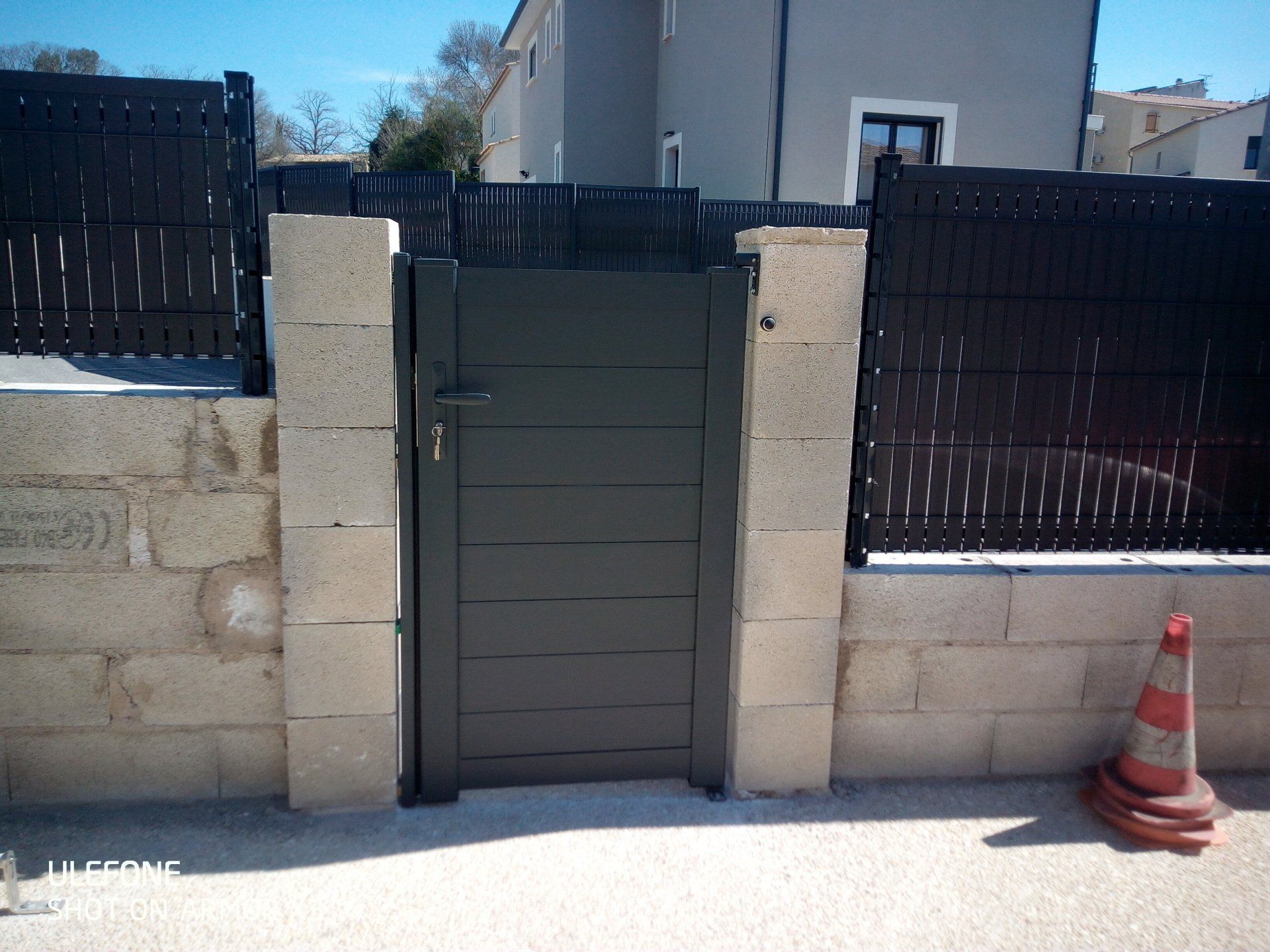 Un portillon piétonnier moderne à lattes horizontales gris foncé, encadré par deux piliers en blocs de béton non enduits.