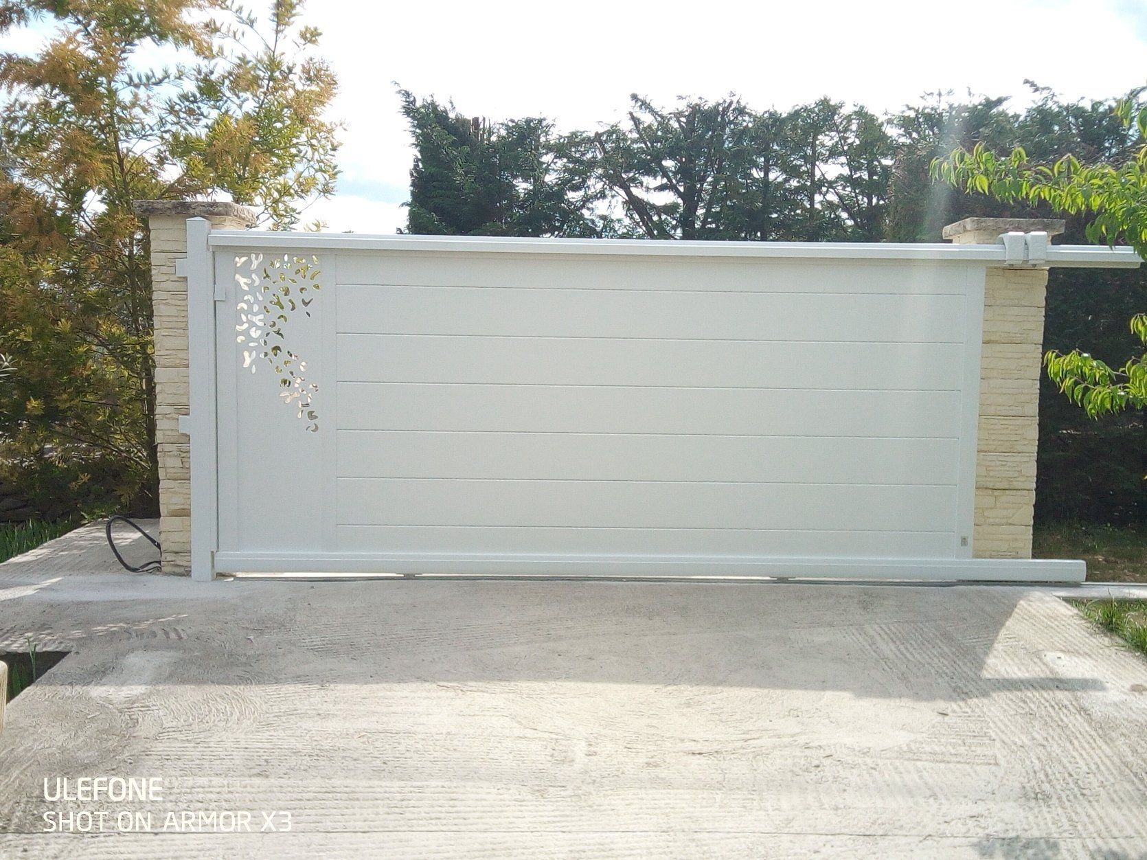 Un portail coulissant blanc pour allée, orné d'un motif floral ajouré, installé entre deux piliers en pierre.
