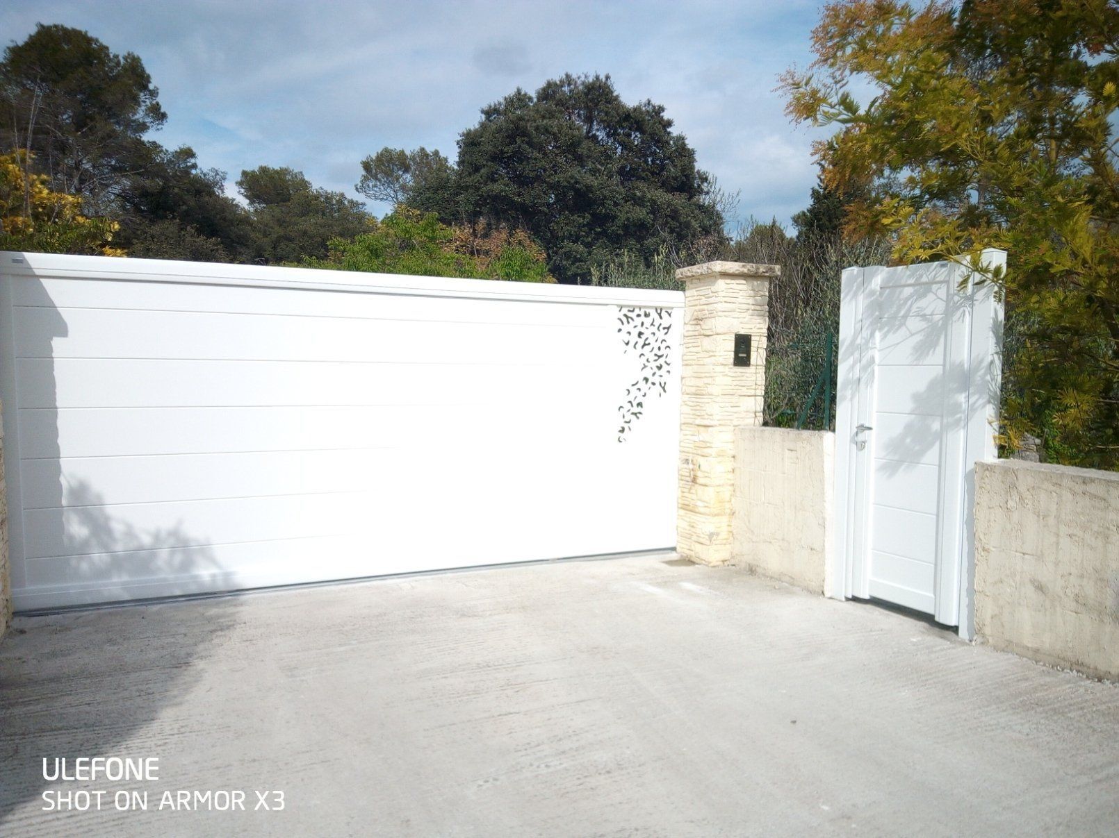 Un portail blanc et une porte piétonne assortie, fixés à un pilier de pierre, se détachent sur un fond d'arbres.