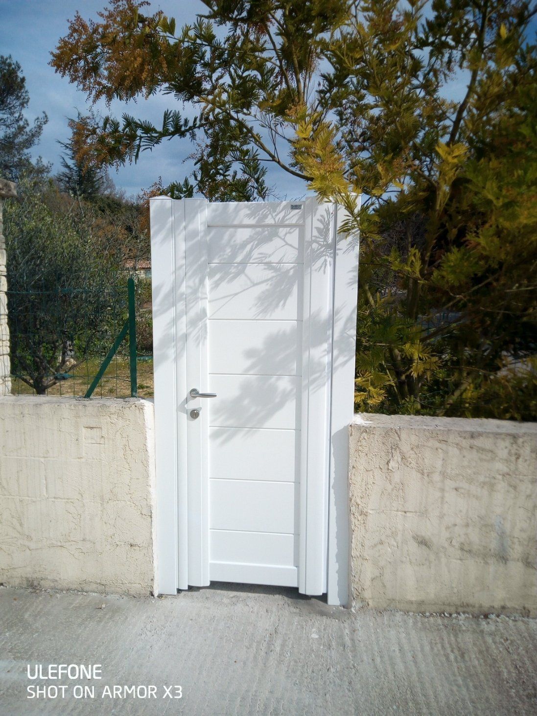 Une porte blanche à panneaux horizontaux et à poignée métallique est encastrée dans un mur de pierre beige, à l'extérieur, sous les arbres.