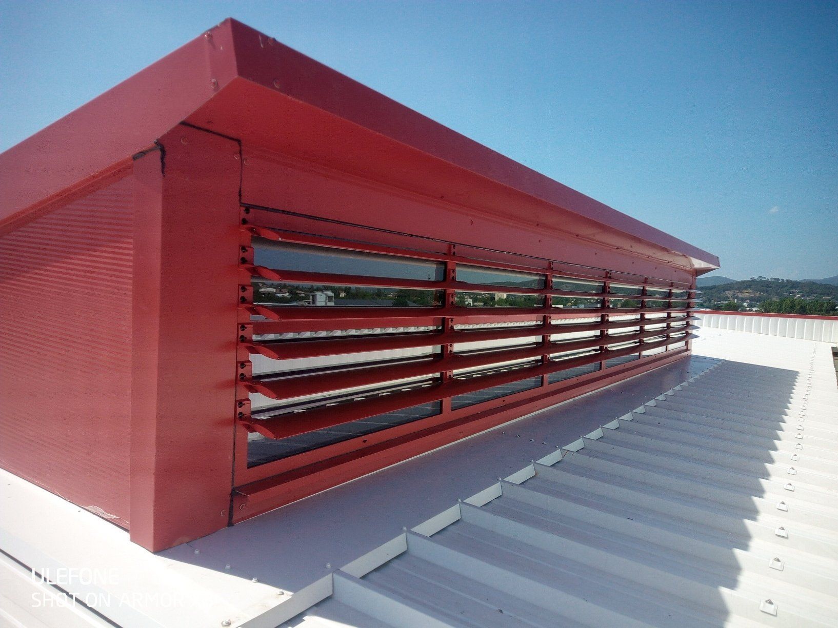 Un lanterneau rouge en métal à persiennes horizontales est installé sur un toit en tôle ondulée blanche, sur fond de ciel bleu.