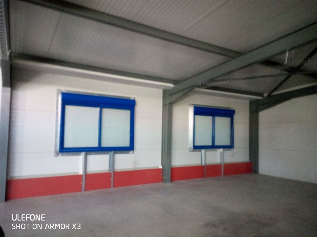 Vue intérieure d'un espace industriel avec des murs blancs, un socle rouge, deux fenêtres à cadre bleu et des poutres métalliques au plafond.