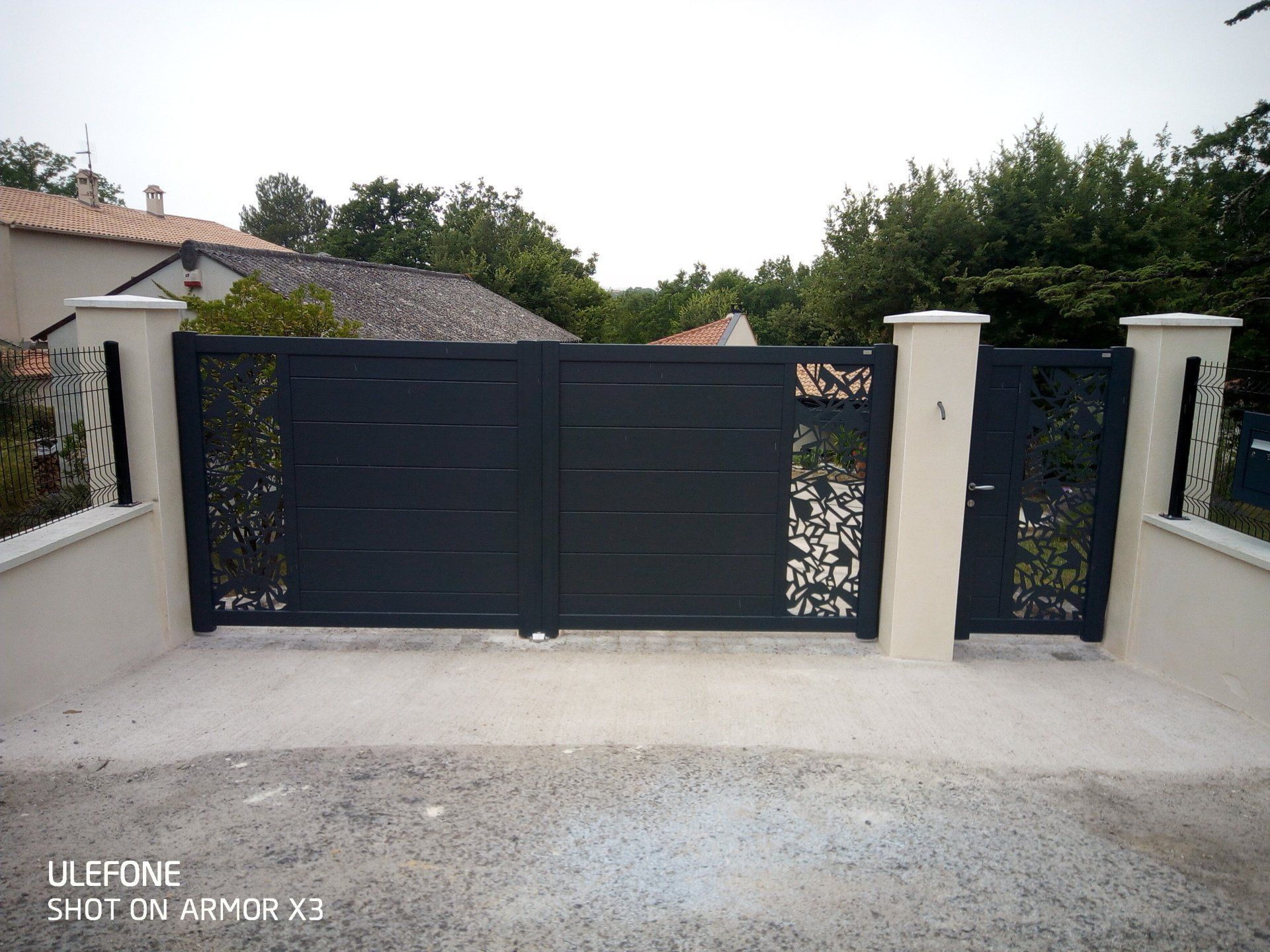 Un portail d'allée moderne noir orné de motifs complexes découpés au laser, flanqué de piliers beiges et d'un portillon assorti.