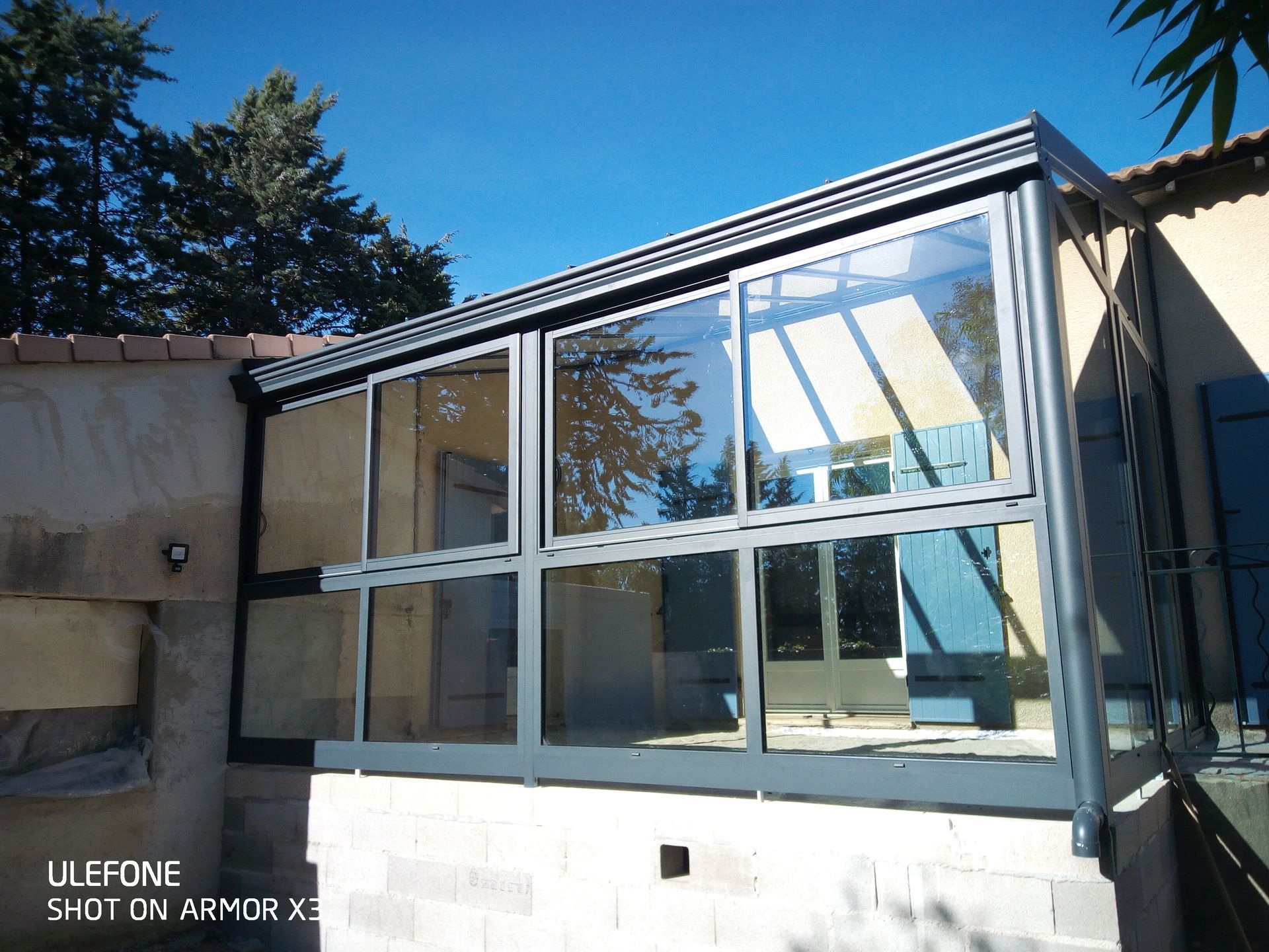 Une structure de patio vitrée avec un cadre métallique gris foncé, fixée sur le côté d'une maison.