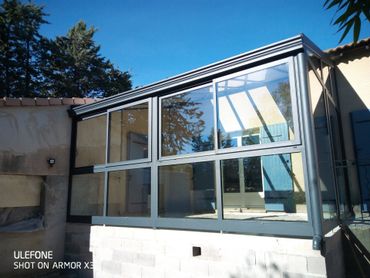Une véranda moderne en aluminium gris, adossée à la façade d'une maison, sous un ciel bleu azur.
