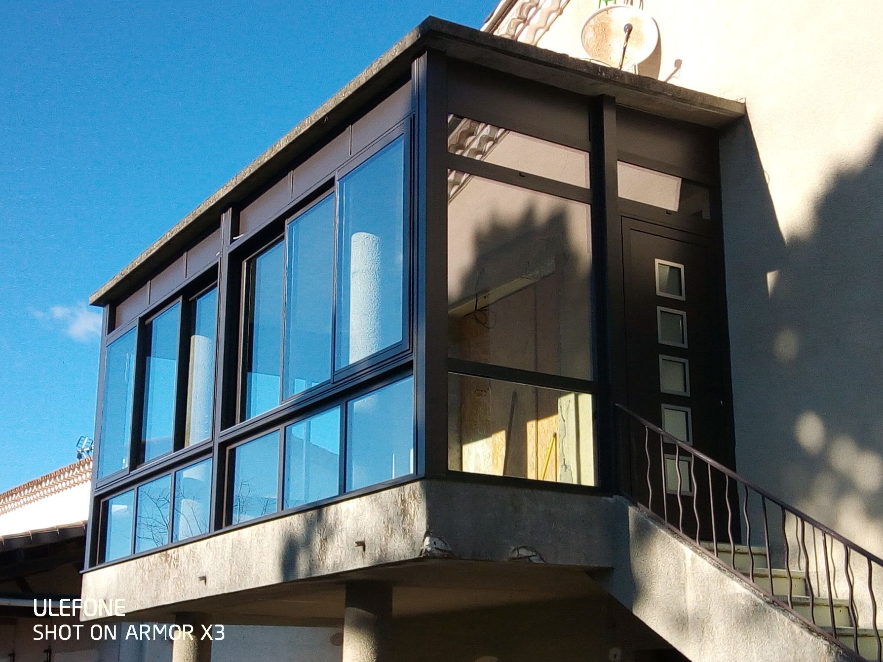Une véranda moderne à cadre sombre, attenante à un bâtiment en pierre, avec des fenêtres coulissantes et une porte latérale.