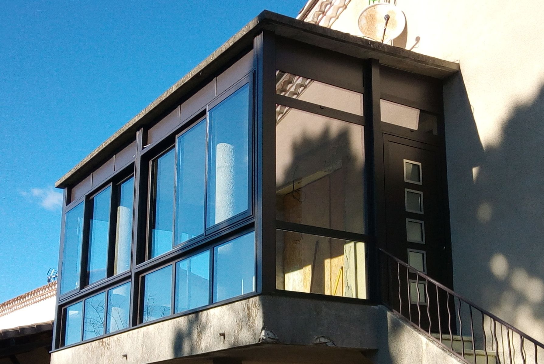 Une structure vitrée à plusieurs panneaux et à cadre sombre, construite sur un balcon en béton et faisant saillie sur le côté d'un bâtiment.