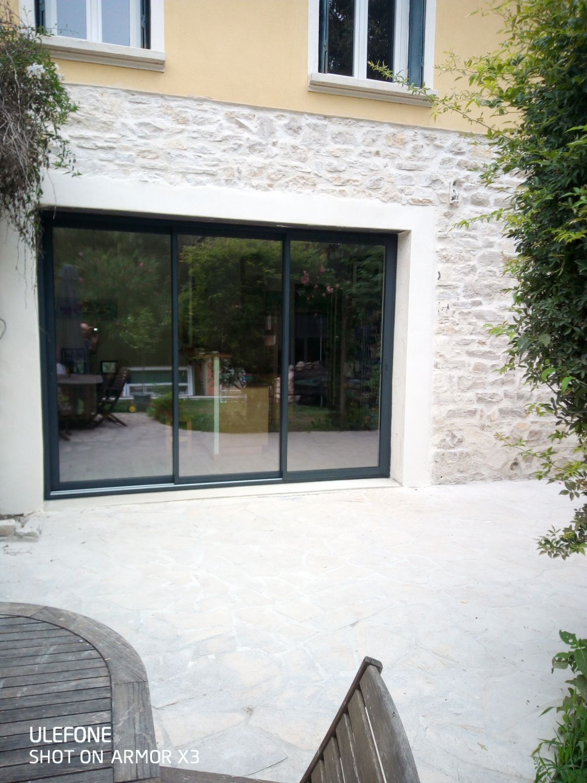 Une grande porte coulissante en verre encastrée dans le mur d'une maison en pierre beige, s'ouvrant sur une terrasse en pierre aménagée avec du mobilier de jardin.