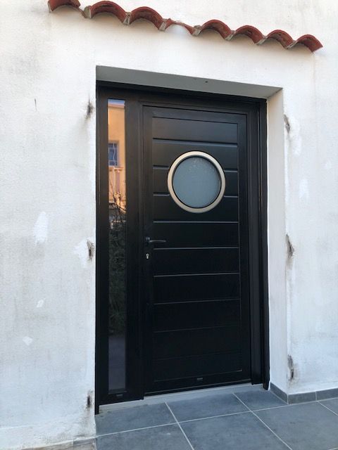 Une porte d'entrée noire à panneaux horizontaux, une fenêtre circulaire et un panneau latéral vitré vertical, le tout encastré dans un mur blanc.