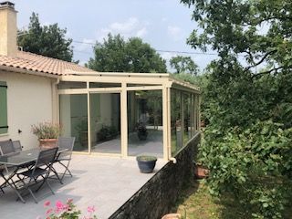 Un patio avec une véranda aux parois de verre beige attenante à une maison, agrémentée de mobilier d'extérieur sur une terrasse carrelée.