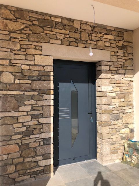 Une porte d'entrée grise avec un panneau de verre vertical encastré dans un mur en maçonnerie de pierres des champs rustiques multicolores.