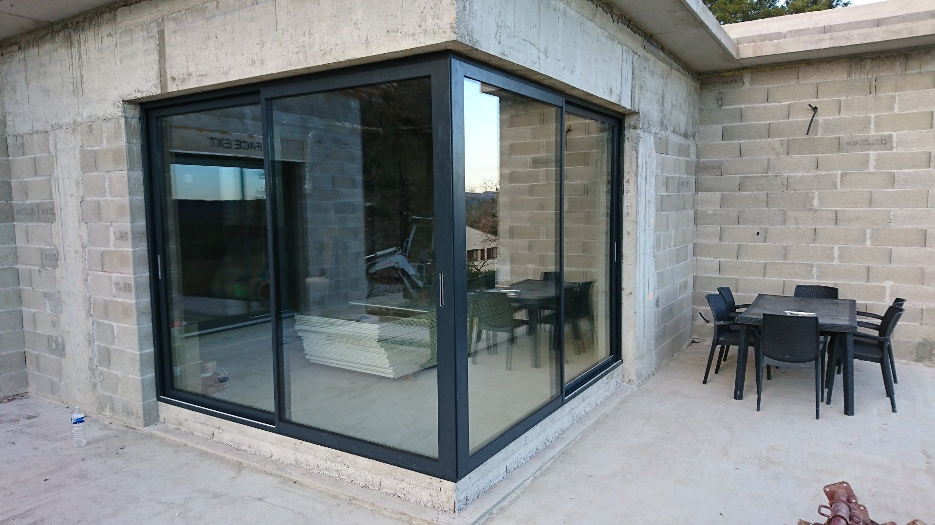 Une porte coulissante d'angle en verre encastrée dans un mur en parpaings de béton gris, avec un ensemble de repas extérieur sur la terrasse à proximité.