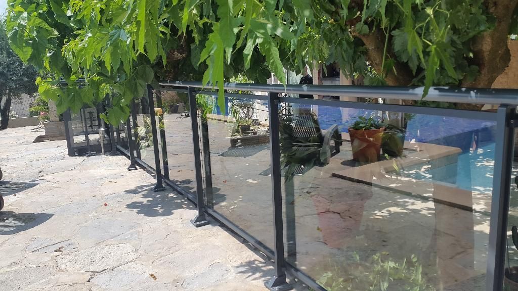 Une balustrade en verre à cadre métallique sombre sépare une terrasse en pierre d'un espace piscine ombragé par des arbres.