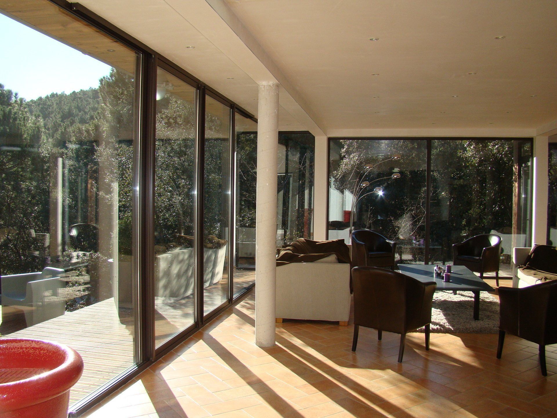 Une pièce intérieure lumineuse et moderne avec des baies vitrées du sol au plafond, un parquet et des chaises sombres donnant sur un jardin extérieur.