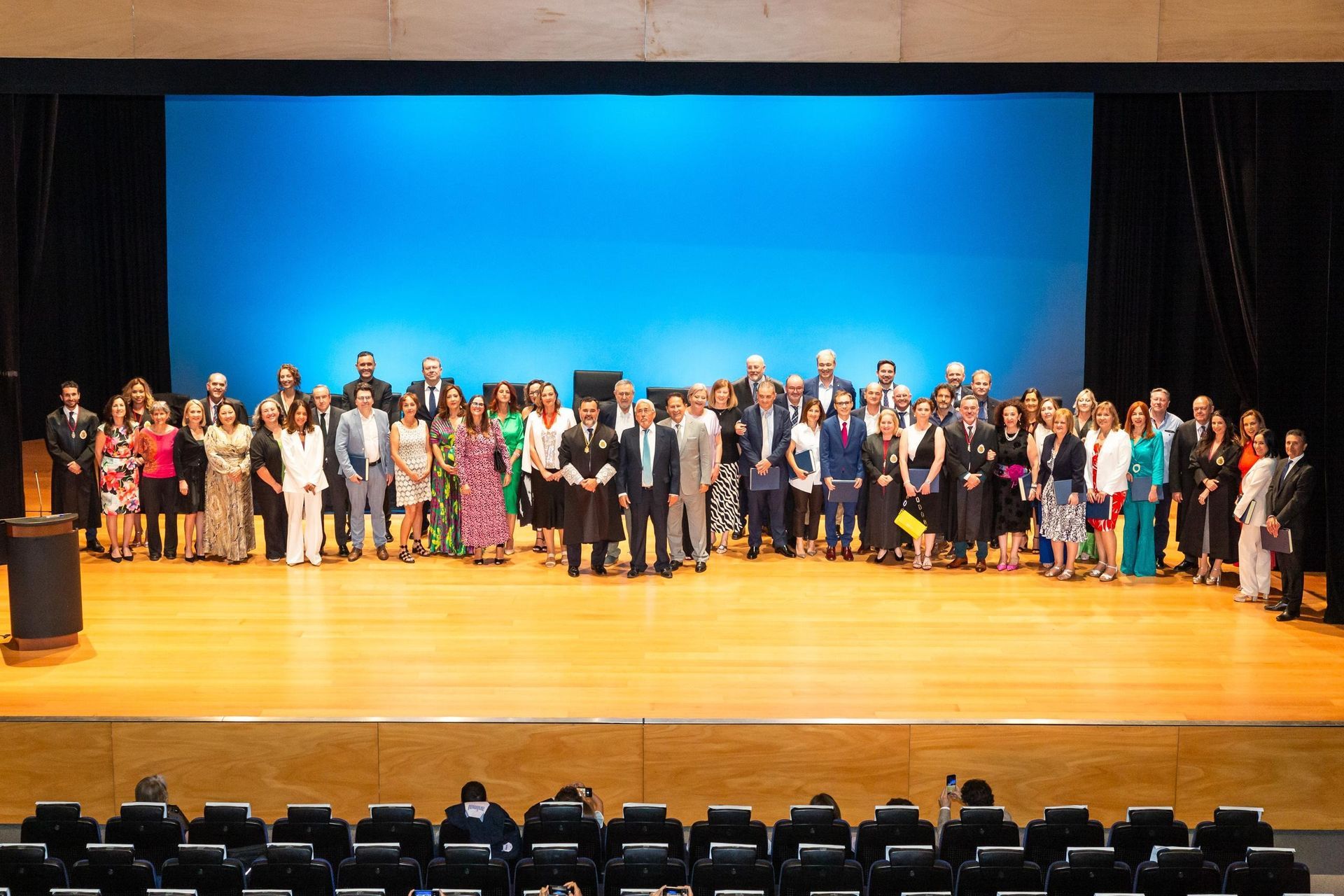 Grupo de personas de pie en un escenario, iluminado con un fondo azul. Vestimenta formal; algunas portan premios o documentos.