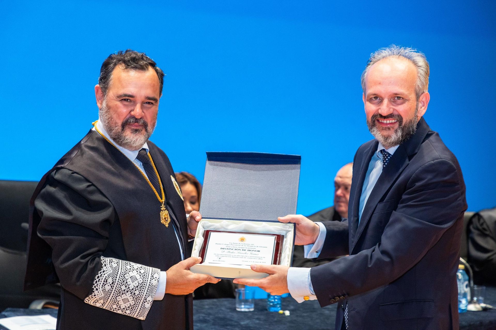 Dos hombres con atuendo formal intercambian una placa de reconocimiento. Uno con túnica negra y el otro con traje; fondo azul.
