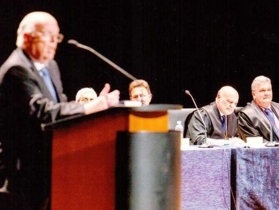 Un hombre habla desde un podio; jueces con togas negras se sientan detrás de él, algunos mirando hacia abajo. Escenario formal, escenario iluminado.