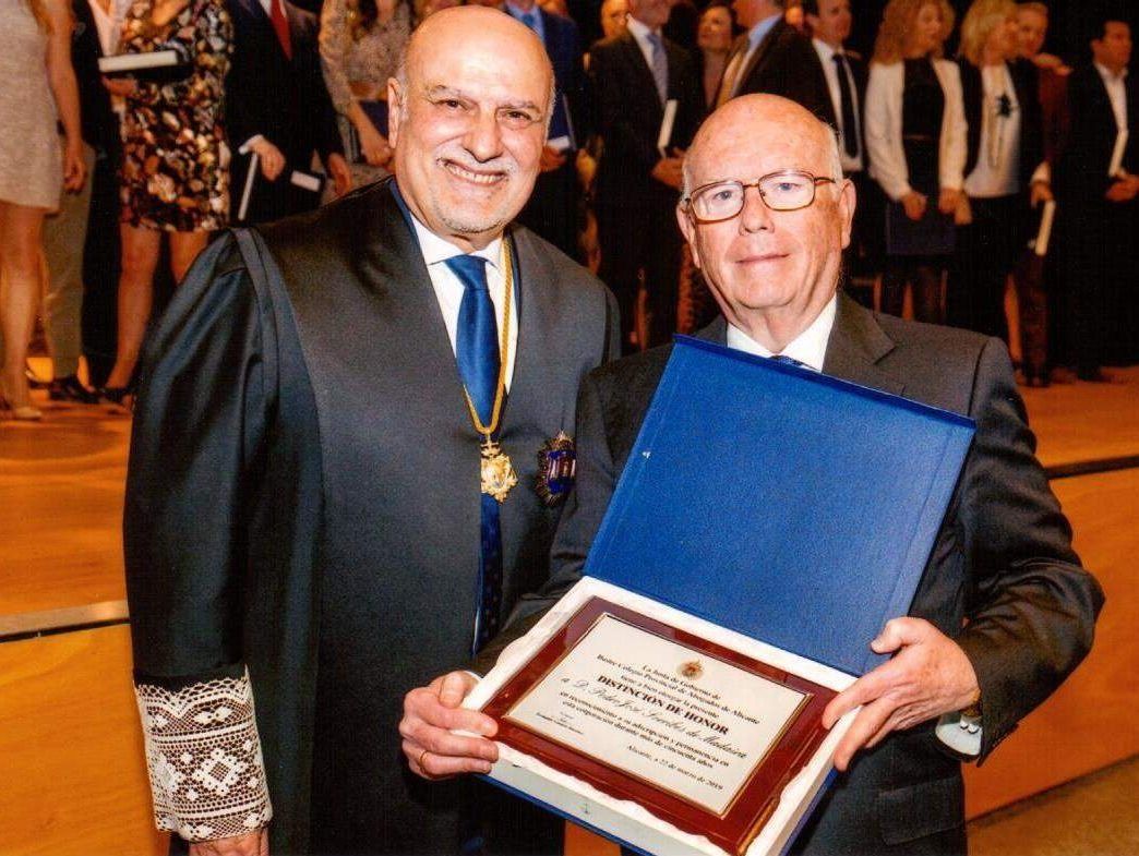Dos hombres mayores con atuendo formal; uno con toga judicial, el otro con una placa. Sonríen en un evento, probablemente una ceremonia de premiación.