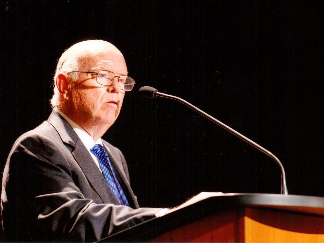 Un hombre con gafas habla en un podio con un micrófono; lleva un traje oscuro y corbata sobre un fondo negro.