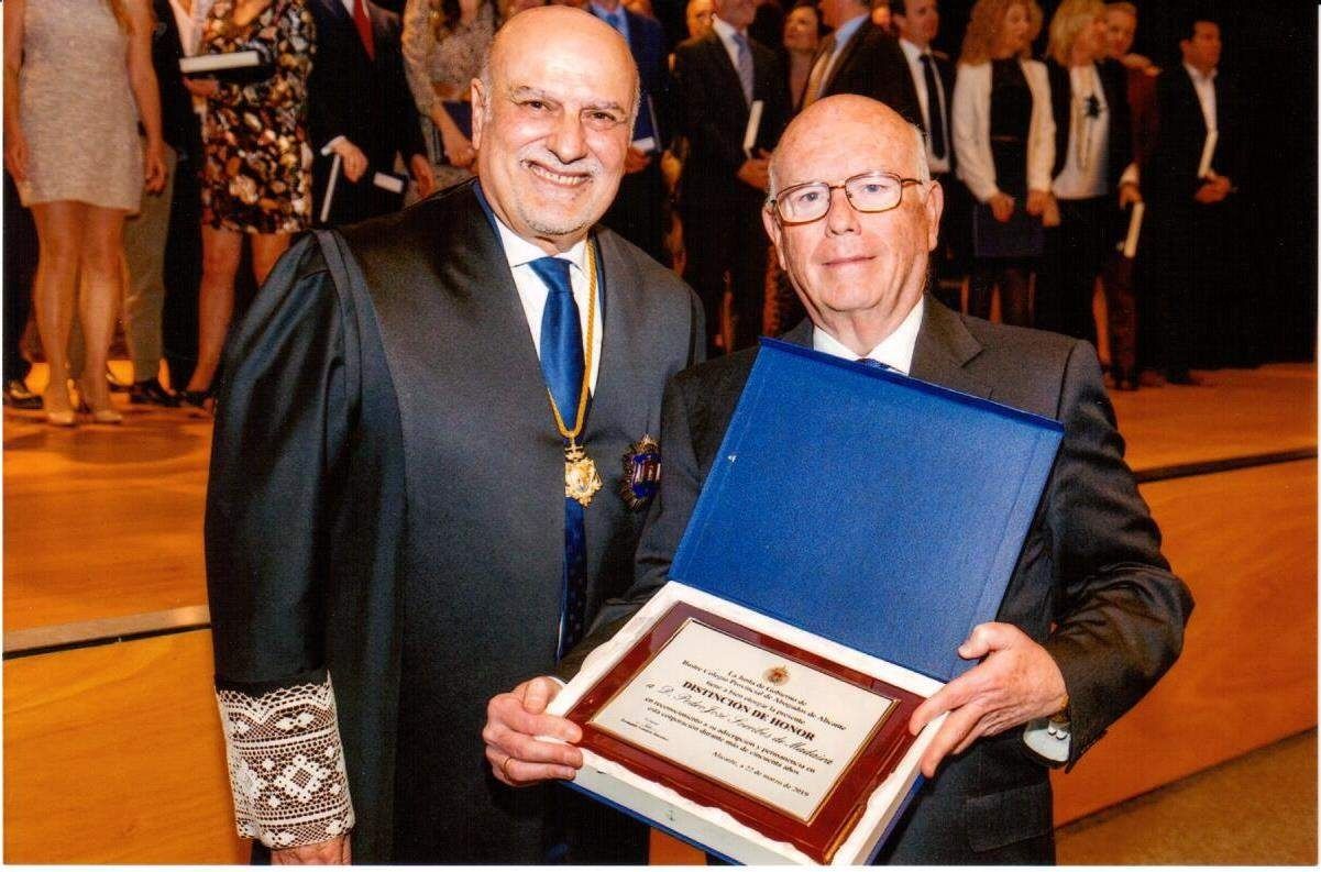 Dos hombres de traje, uno de ellos con togas judiciales, posando y sosteniendo una placa de premio en un espacio interior, probablemente en una ceremonia.