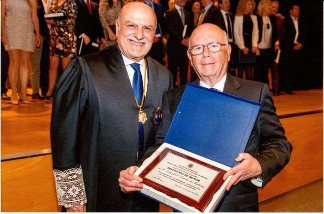 Dos hombres de traje, uno de ellos con togas judiciales, posando y sosteniendo una placa de premio en un espacio interior, probablemente en una ceremonia.