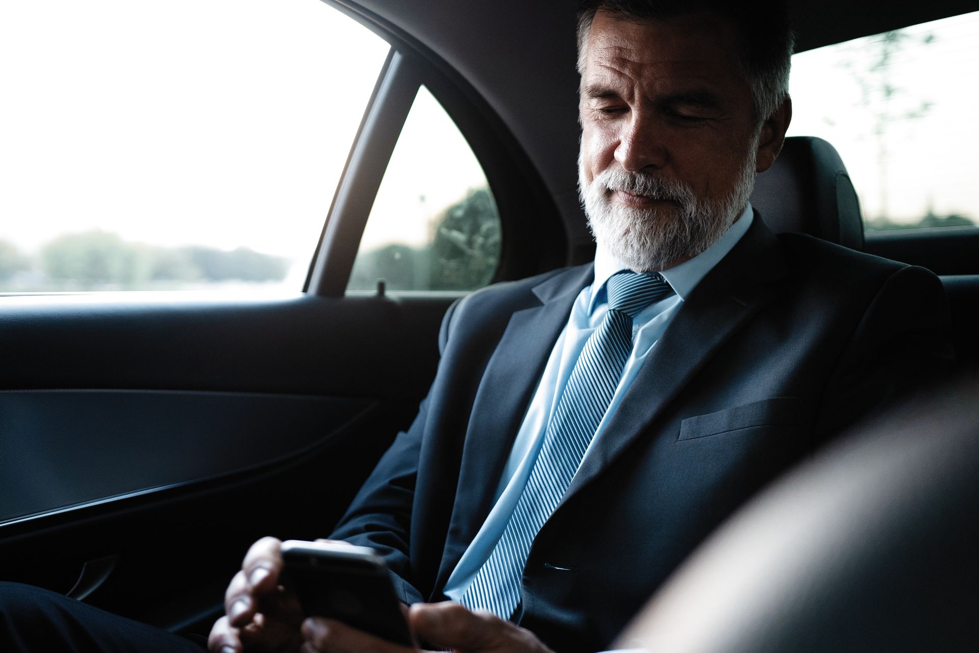 Un homme en costume assis dans une voiture, regardant son téléphone près de la fenêtre
