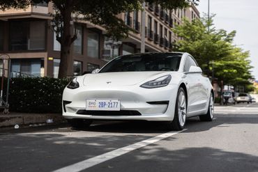 Berline Tesla blanche garée dans une rue en ville, à côté d’immeubles d’habitation et d’arbres.
