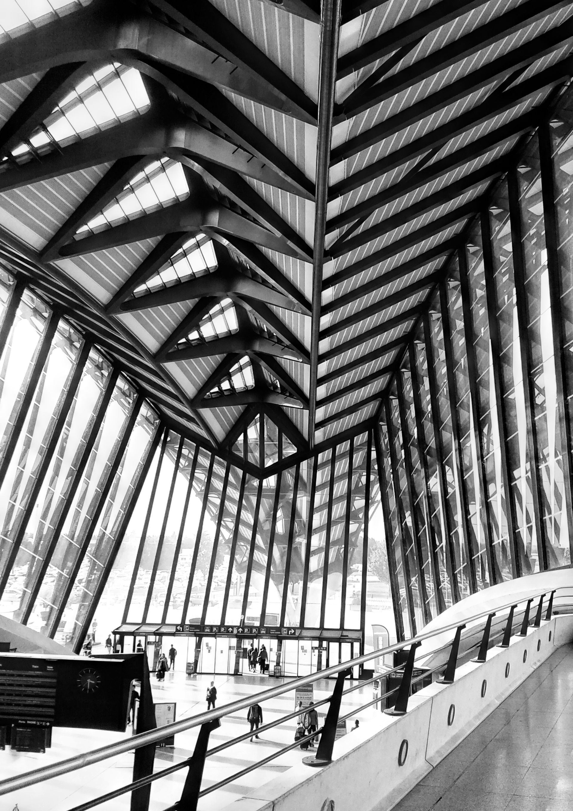 Vue en noir et blanc d’un atrium moderne en verre, avec des poutres inclinées et une passerelle incurvée