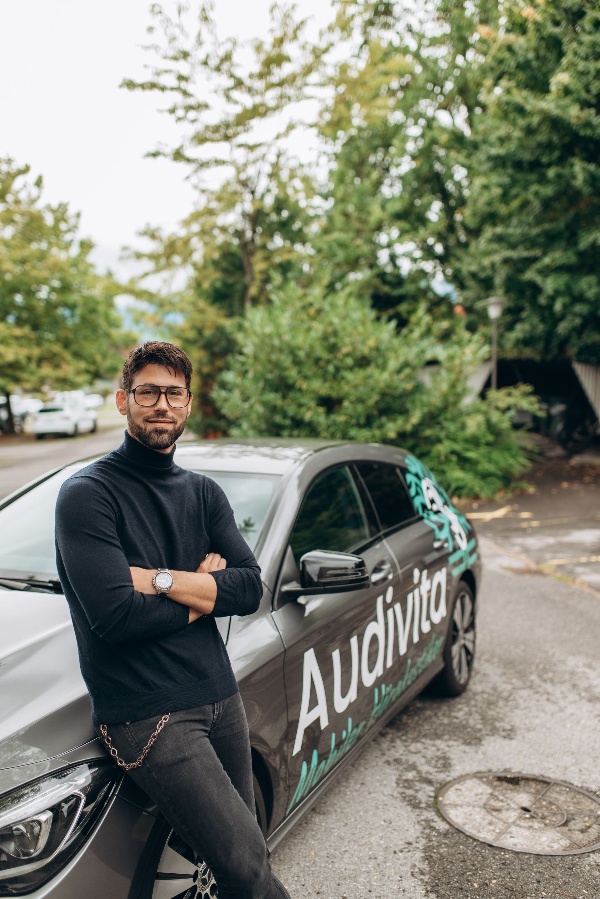 Ein Mann lehnt an einem grauen Auto mit „Audivita“-Logo. Im Freien, im Hintergrund Bäume.