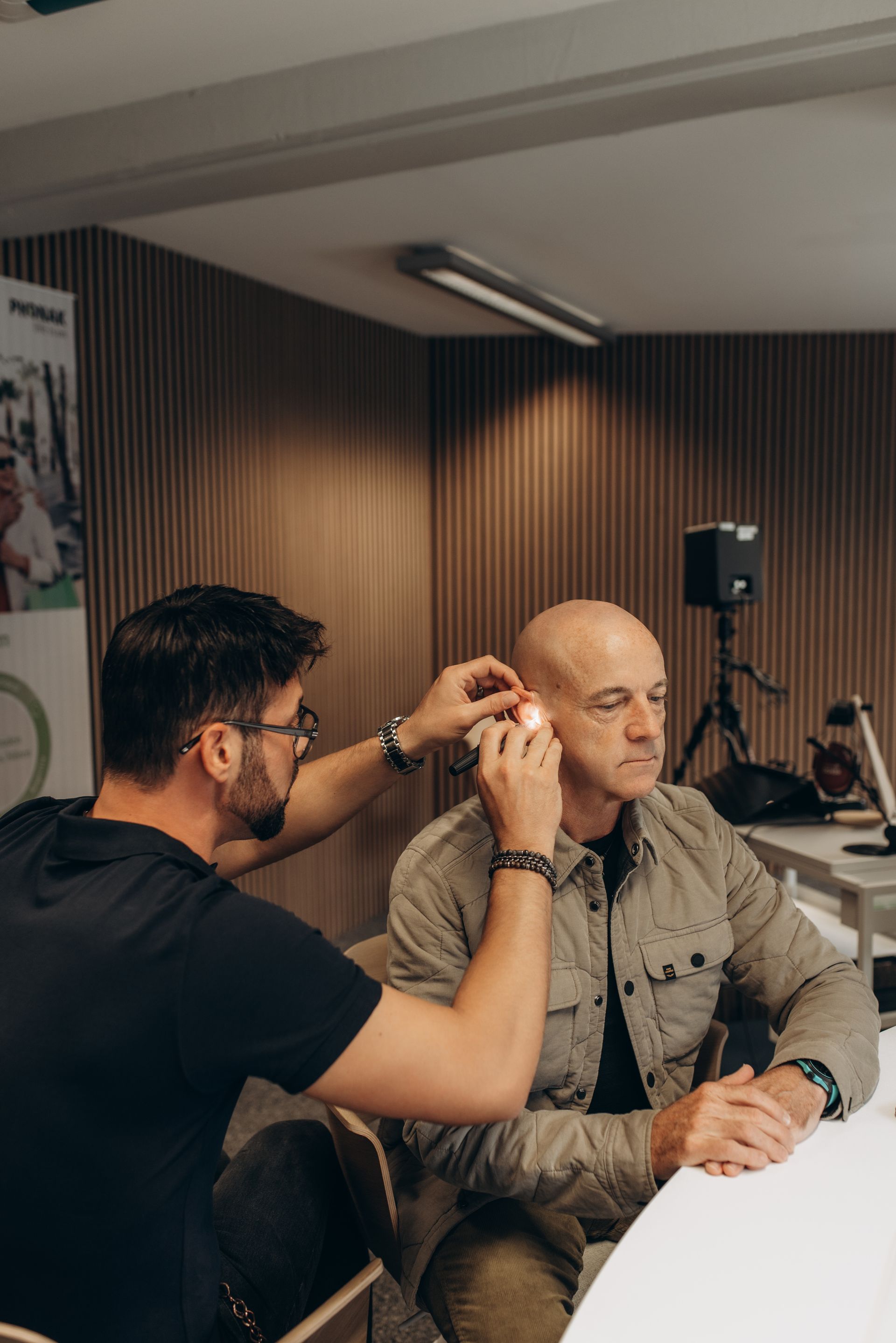 Eine Person unterzieht sich einem Hörtest. Eine andere Person untersucht ihr Ohr mit einem Instrument. Heller Raum.