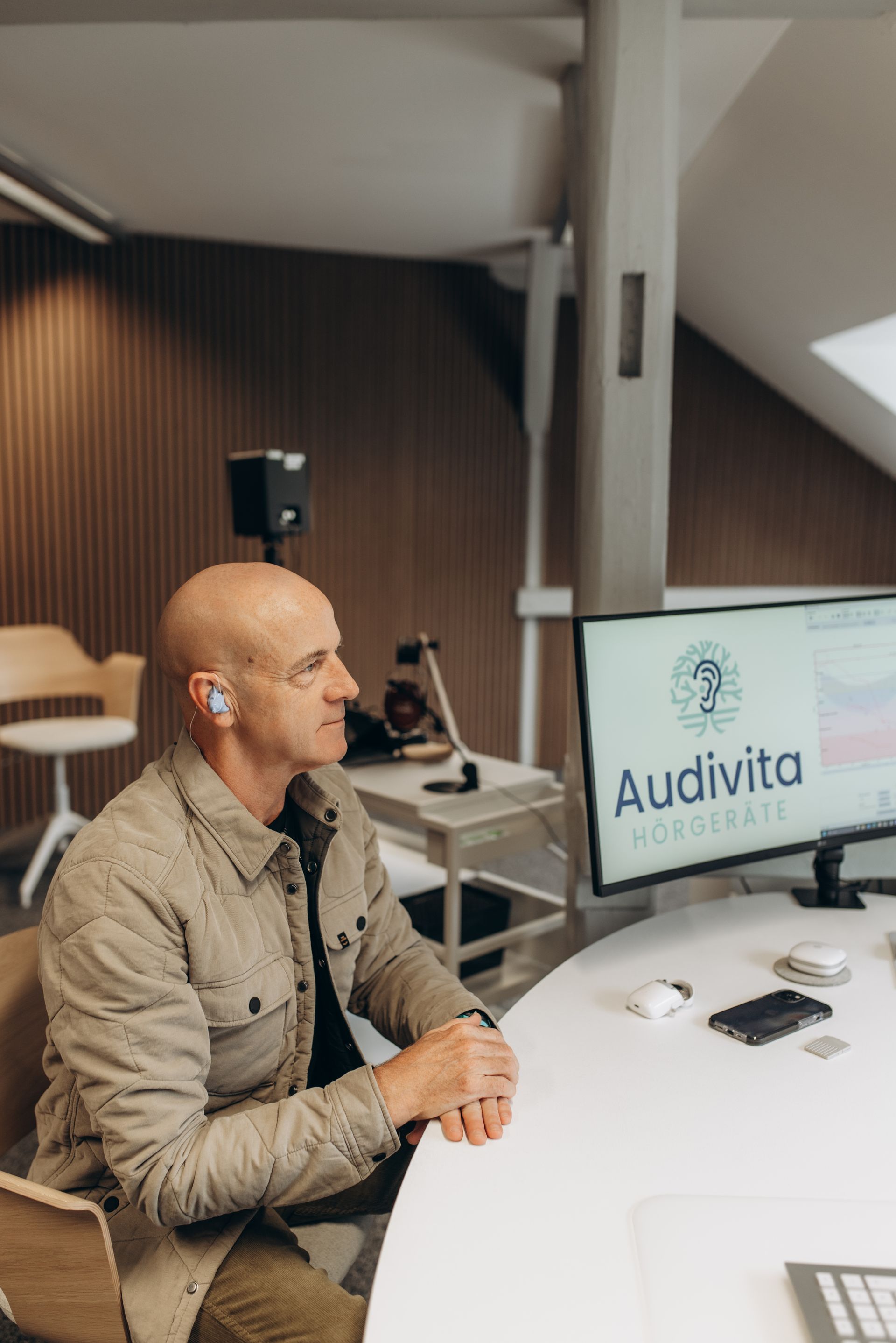 Ein Mann mit Brille sitzt an einem Tisch und wird interviewt; ein anderer Mann benutzt ein Tablet. Hintergrund: Holzvertäfelung.