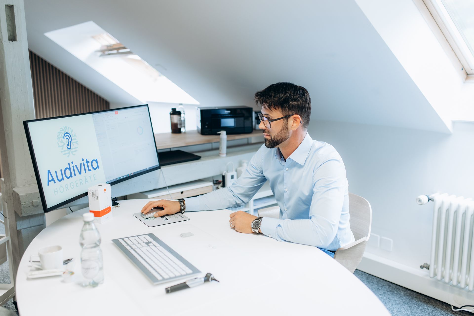 Mann an einem Schreibtisch mit zwei Monitoren; auf einem davon das Audiovita-Logo. Weisser Schreibtisch, hellblaues Hemd, Brille, Inneneinrichtung.