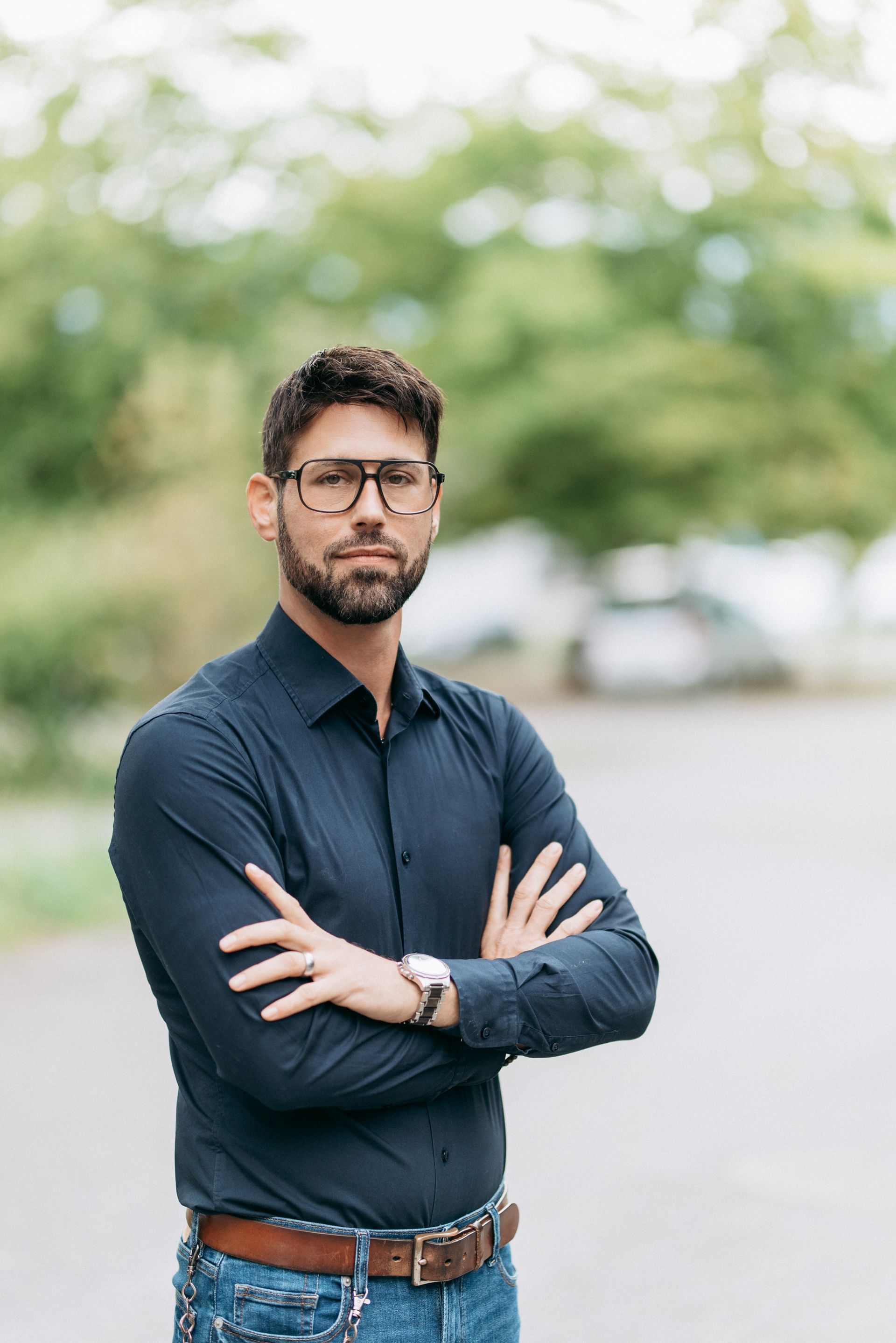 Eine Frau und ein Mann sitzen in einem Büro und posieren. Der Mann trägt eine Brille und ein dunkles Hemd. Die Frau trägt einen Blazer.