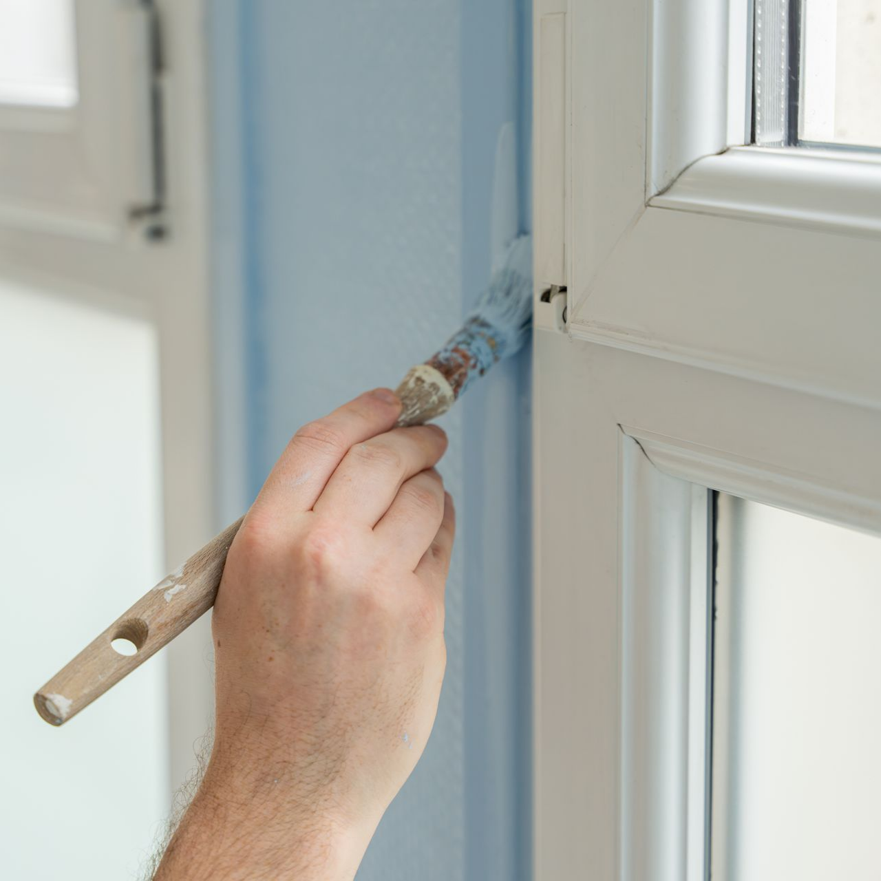 Une main utilise un pinceau pour peindre un mur bleu pâle le long du bord d'un cadre de fenêtre blanc.