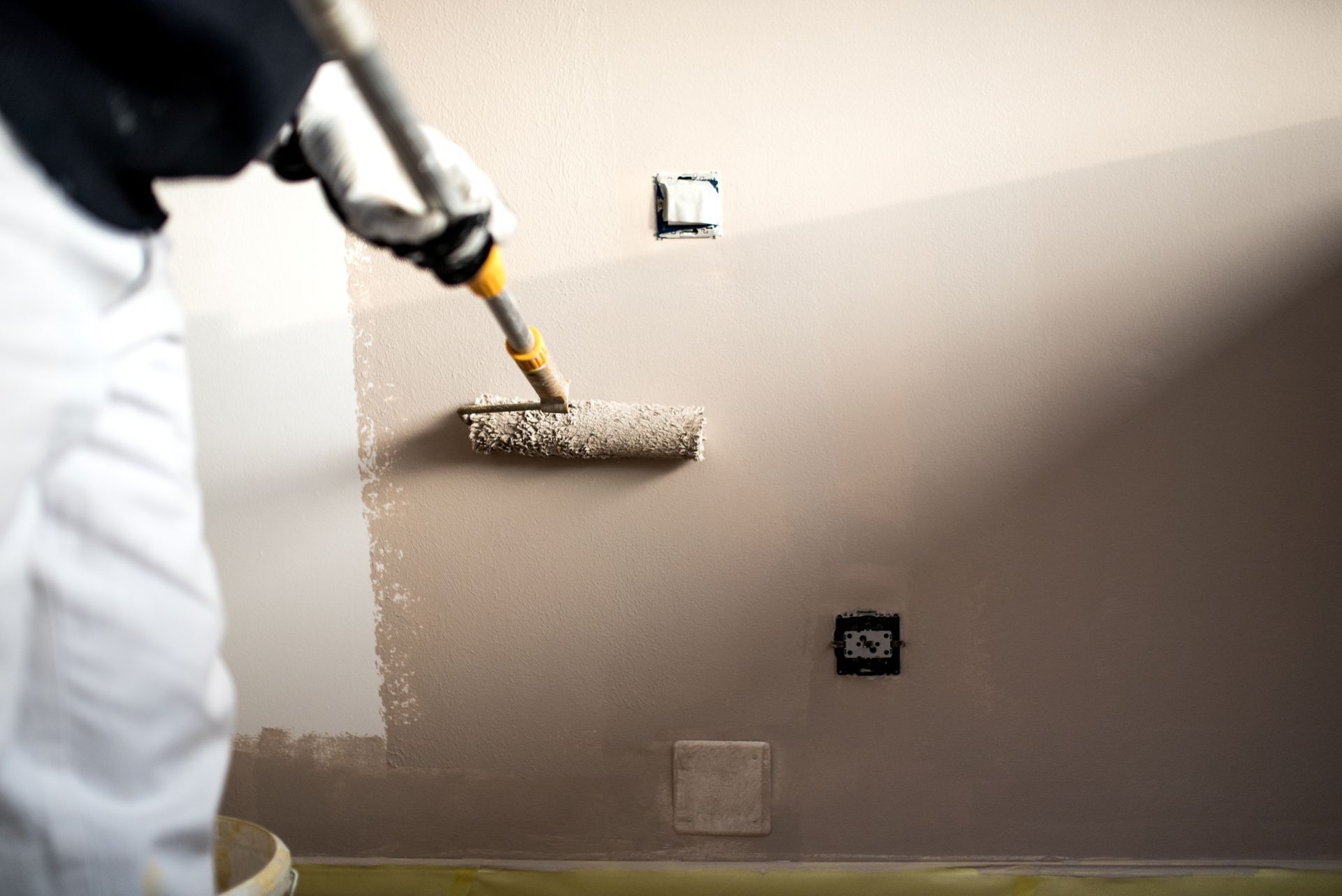 Une personne en tenue de travail blanche utilise un rouleau à peinture pour appliquer une peinture gris-brun sur un mur comportant des prises électriques.