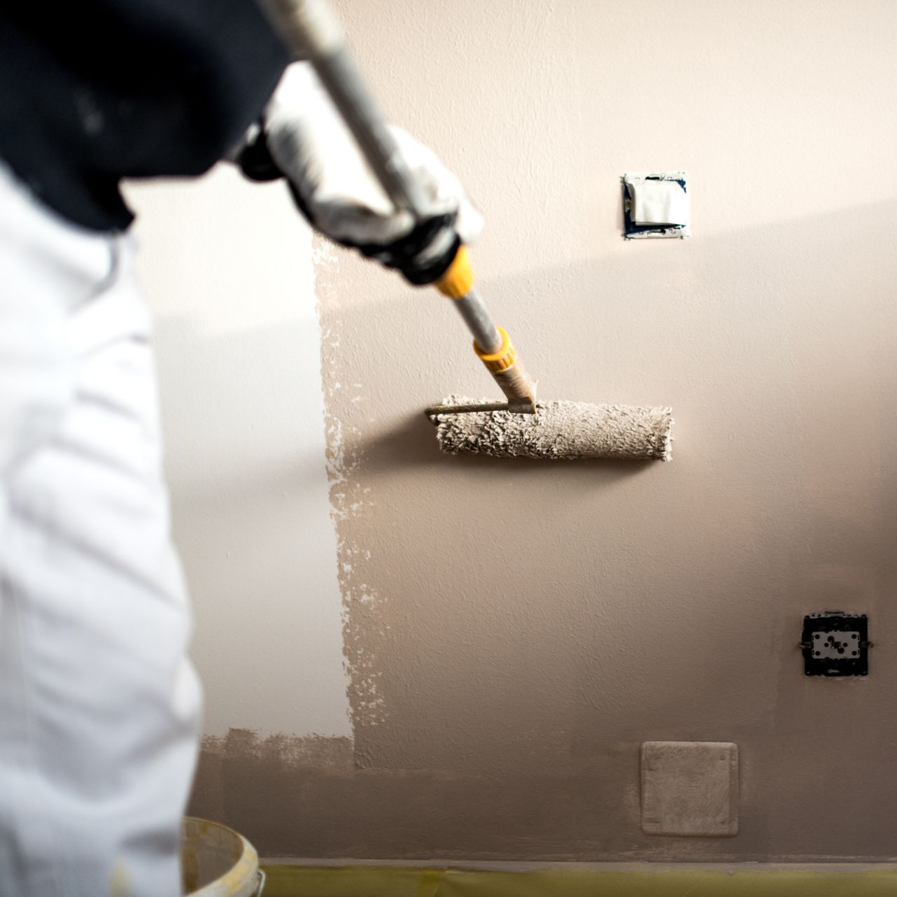 Une personne en pantalon blanc utilise un rouleau à peinture fixé à une perche télescopique pour appliquer de la peinture brun clair sur un mur intérieur.