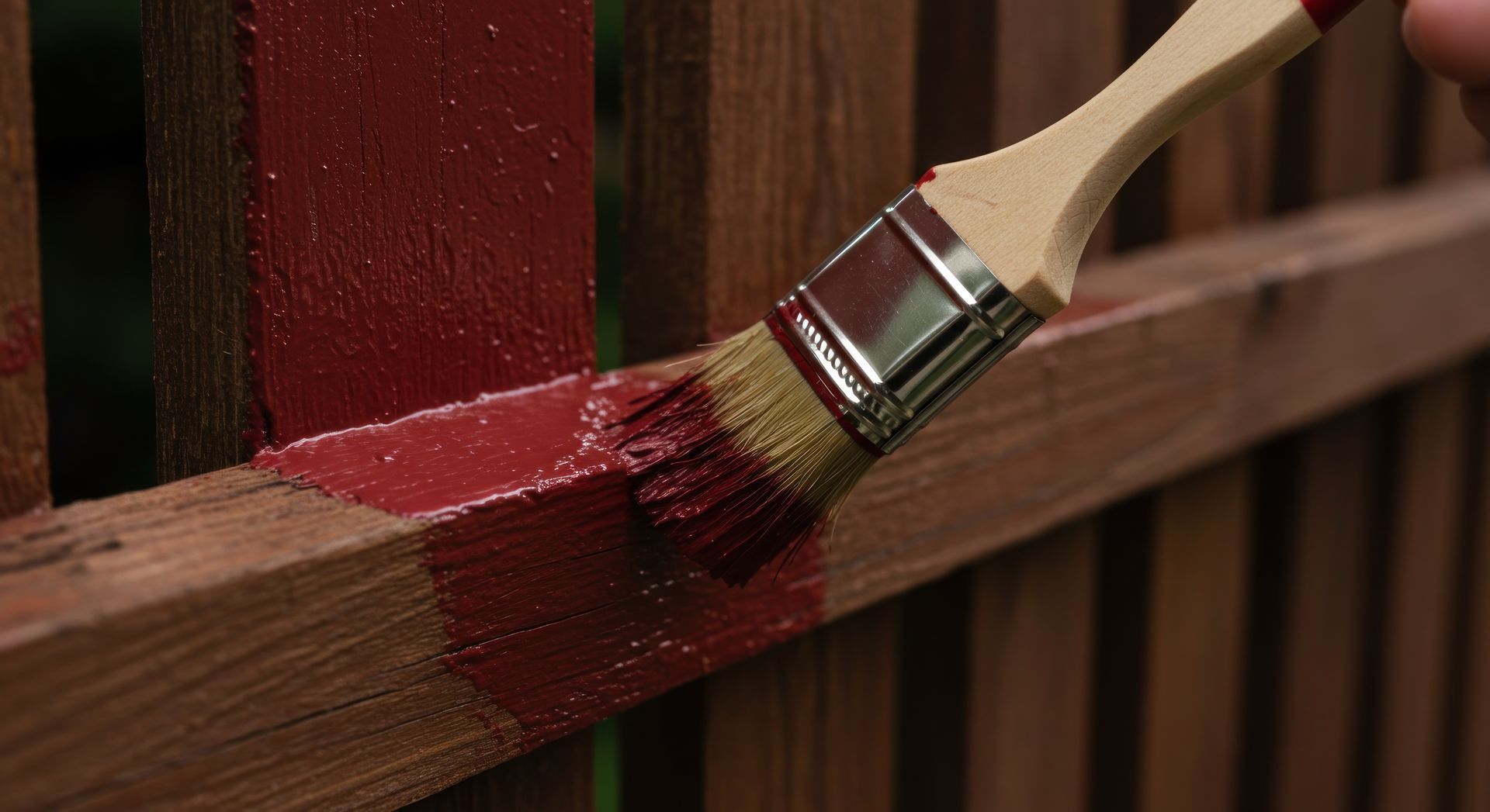Une main tient un pinceau à manche en bois, appliquant une peinture rouge foncé sur les poutres horizontales et verticales d'une clôture en bois.