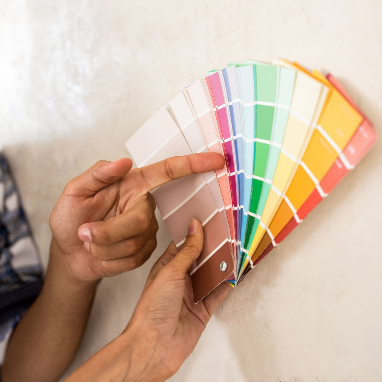Des mains tiennent et désignent un nuancier de couleurs devant un mur uni.