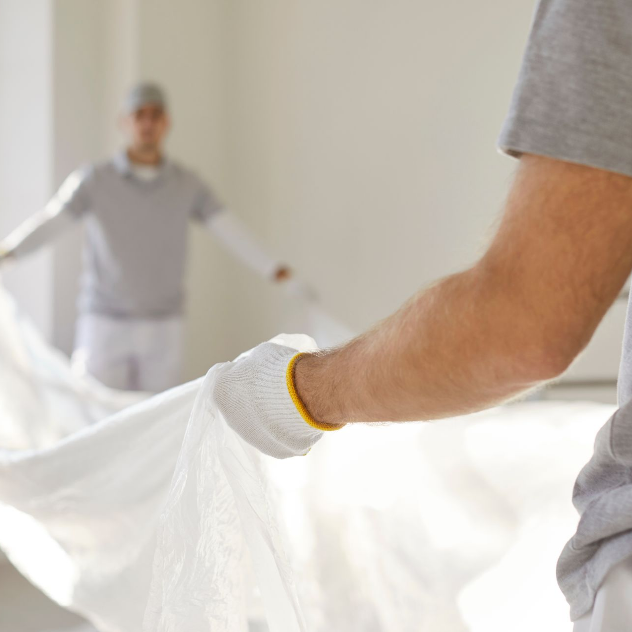 Deux personnes vêtues de chemises grises et de gants de travail déplient ensemble une grande bâche blanche dans une pièce vide.