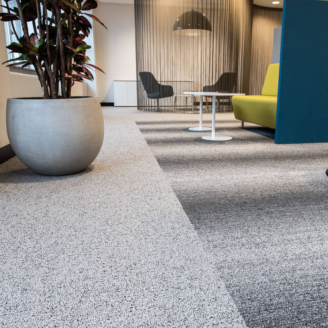 Un coin salon de bureau moderne avec une grande plante en pot, un canapé jaune et de petites tables d'appoint blanches sur une moquette grise mouchetée.