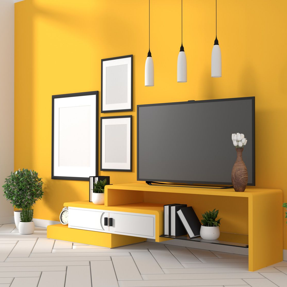Un mur jaune est orné de trois affiches encadrées, d'une télévision sur un meuble TV jaune, de suspensions blanches et de petites plantes.