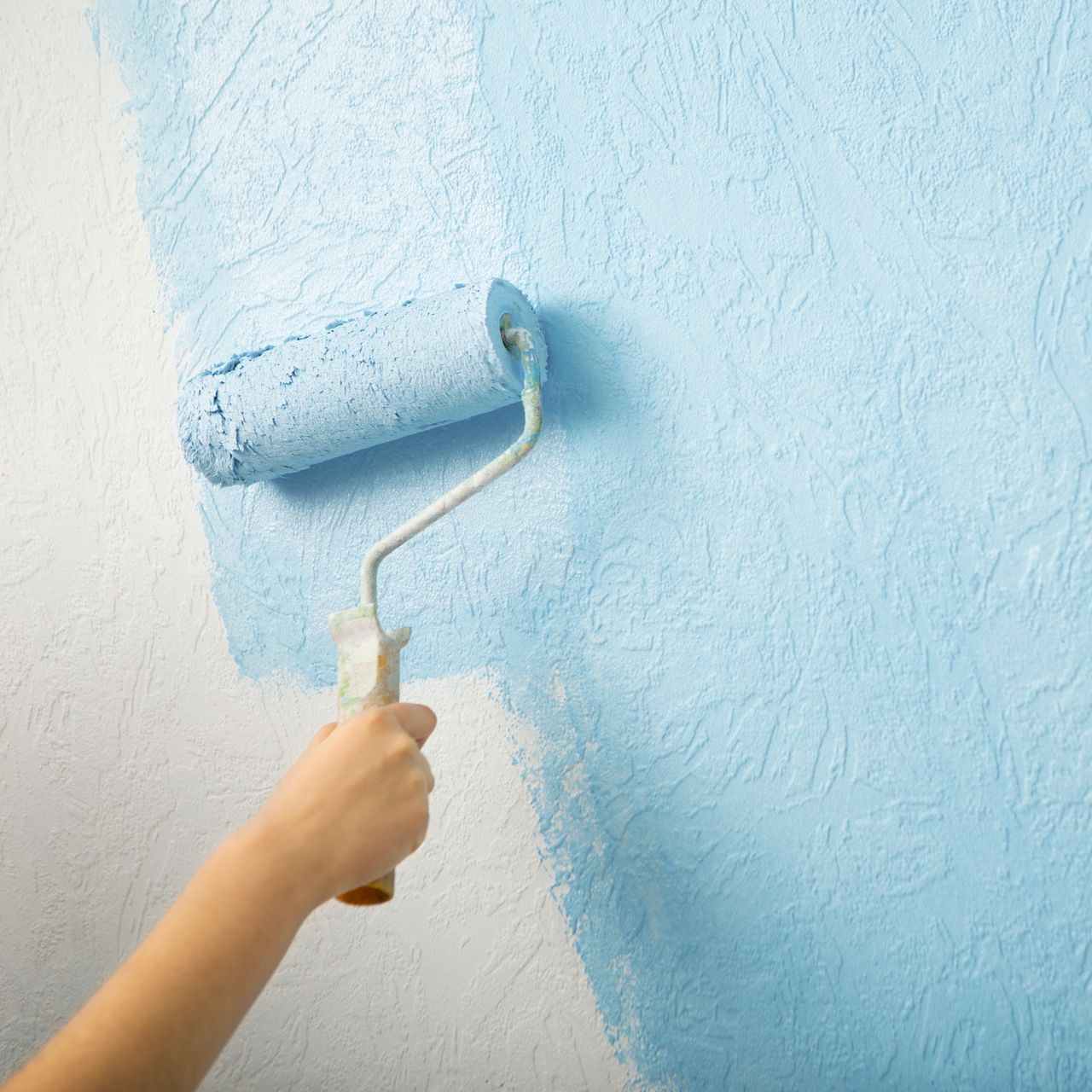 Une main utilise un rouleau à peinture pour appliquer de la peinture bleu clair sur un mur blanc texturé.
