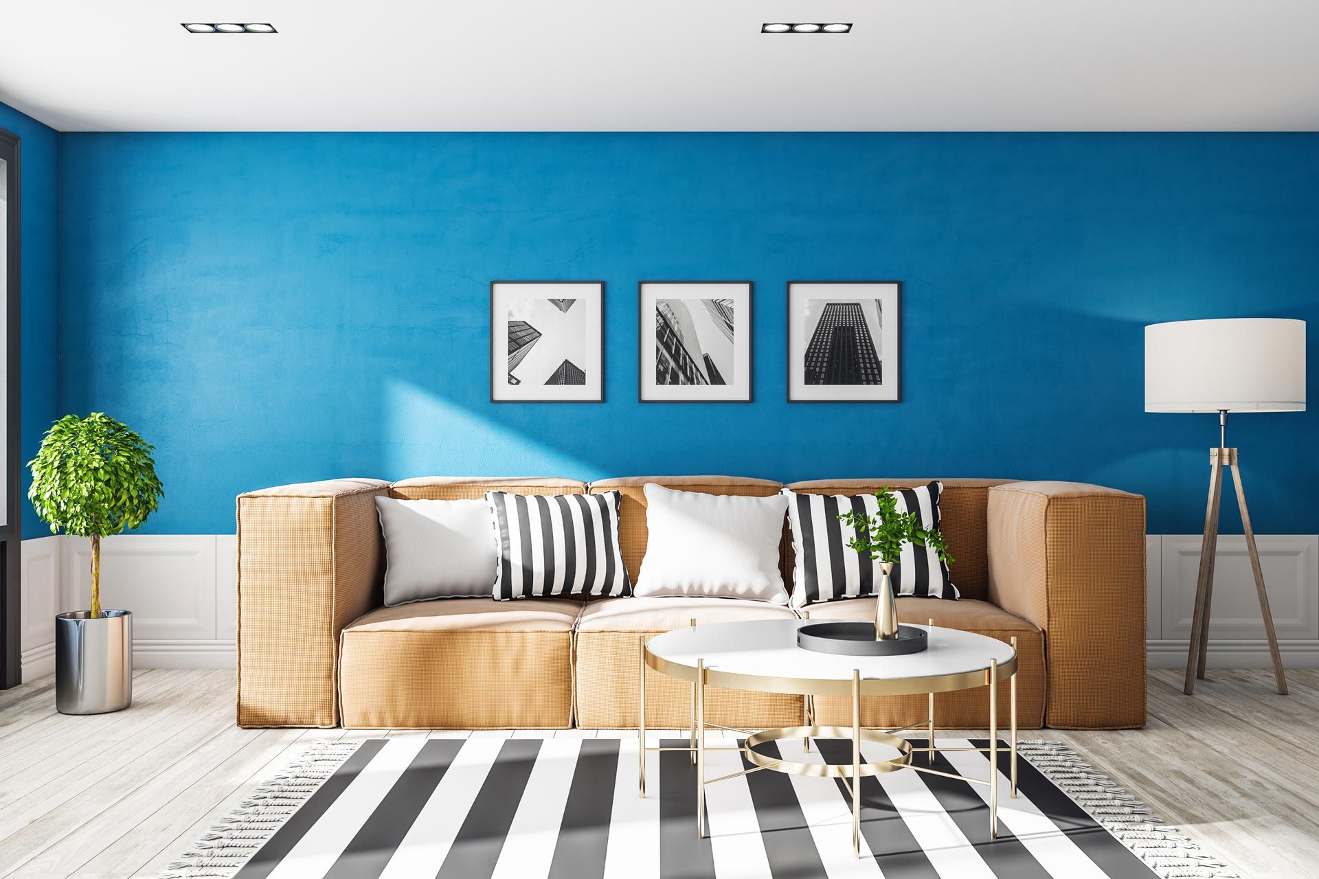 Un canapé beige orné de coussins rayés et blancs est placé devant un mur bleu où sont accrochés trois estampes encadrées et un lampadaire.