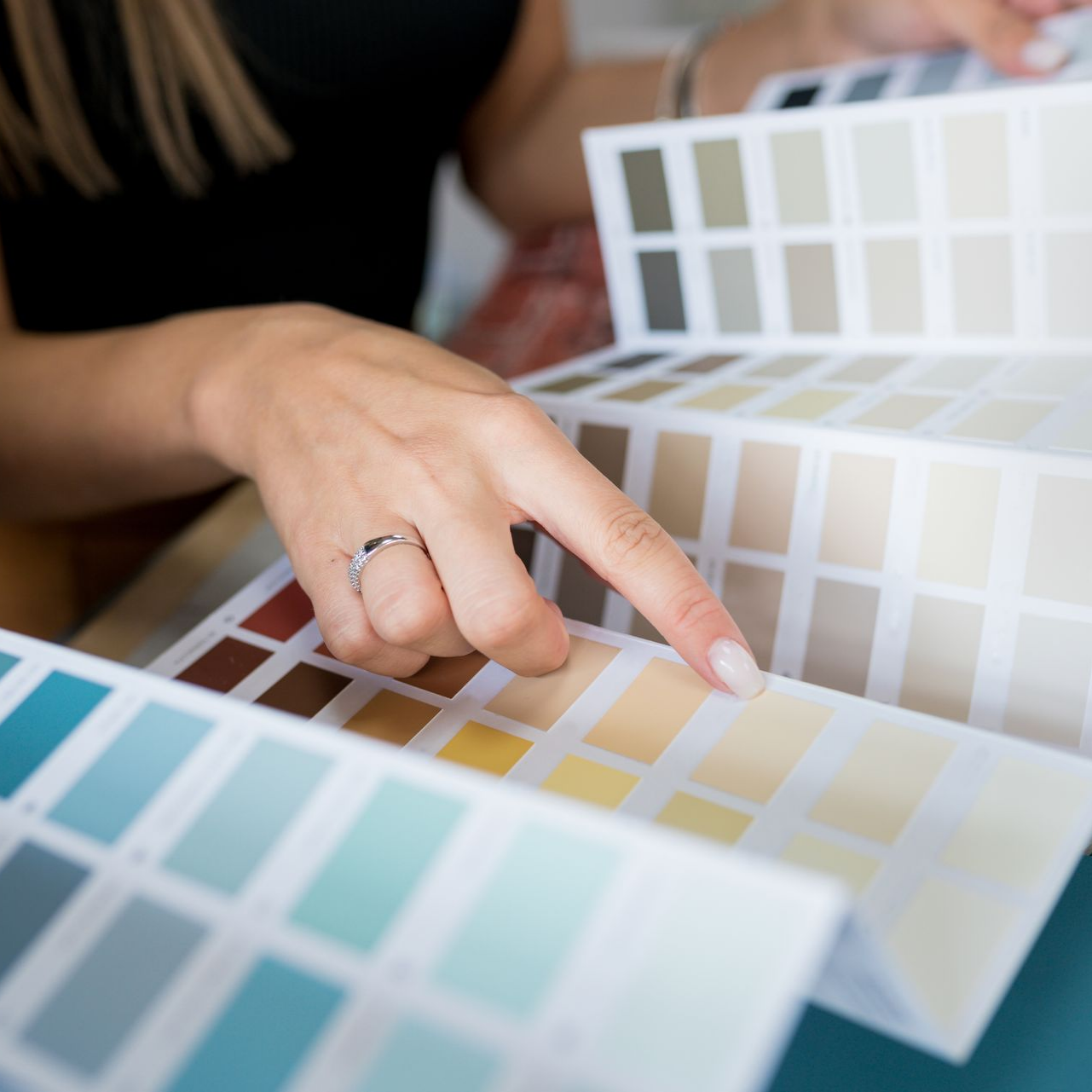 Une main désigne un échantillon de couleur crème claire sur un nuancier de peinture, entouré d'autres palettes dans des tons bleus et neutres.